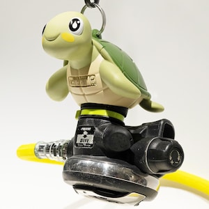 Puede incluir: Un regulador de buceo en forma de tortuga verde y amarillo con una boquilla negra y plateada y una manguera amarilla. El regulador tiene el texto "Poldive Dive" impreso en él.
