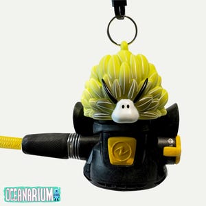 Könnte beinhalten: Ein schwarz-gelber Tauchregler mit einem dekorativen gelb-weißen Design auf der Oberseite. Der Regler hat einen gelben Schlauch und einen Metallring zur Befestigung. Das Wort "Oceanarium" ist in Türkis gedruckt.