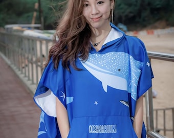 May include: Blue hooded poncho with a whale shark design. The poncho features a large whale shark illustration on the front and a pocket with the text "OCEANARIUM". The poncho is made of a soft, lightweight material, perfect for beach or pool use.