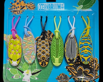 May include: A package of six oval-shaped keychains with colorful designs and matching elastic loops. The keychains feature various patterns in yellow, green, black, blue, and white. The package has the text "OCEANARIUM".
