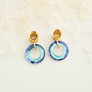 Könnte beinhalten: Ein Paar goldfarbene Ohrringe mit kreisförmigem, offenem Design. Die Kreise haben einen Farbverlauf von Blau und Türkis. Die Ohrringe werden auf einer weißen Oberfläche mit weißen Blumen im Hintergrund präsentiert.