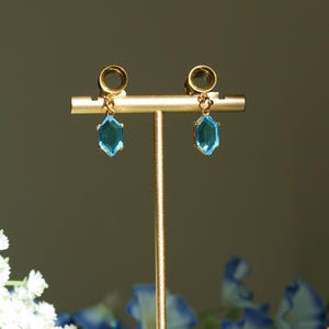 Puede incluir: Un par de pendientes dorados con un colgante geométrico de piedra preciosa azul claro. Los pendientes se exhiben en un soporte dorado, con flores blancas y azules en el fondo. Un accesorio elegante.