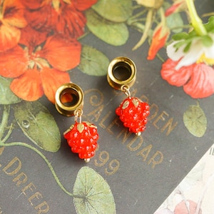 May include: A pair of gold-colored tunnel earrings with dangling red strawberry charms. The earrings are displayed on a dark background with floral and botanical elements, including orange and yellow flowers.