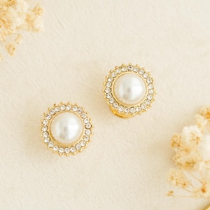 May include: A pair of round stud earrings featuring a central white faux pearl surrounded by a halo of small clear crystals set in a gold-toned metal frame. The circular design highlights the contrast between the smooth pearl and the faceted stones.