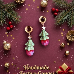 May include: A pair of gold-toned ear gauges featuring dangling charms shaped like green Christmas trees with small silver stars at the top and round red beads at the bottom. The text "Handmade, Festive-Gauges" is printed in gold lettering at the bottom.