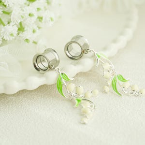 Puede incluir: Un par de expansores de oreja plateados con colgantes de lirios de los valles. Los colgantes presentan capullos de flores blancos y hojas verdes, sobre un fondo blanco suave.