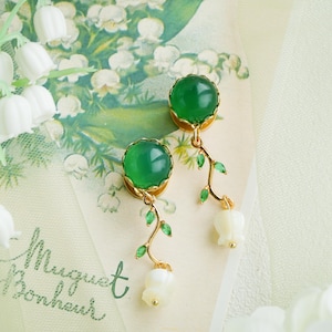 Puede incluir: Un par de pendientes dorados con grandes gemas verdes redondas. Delicadas enredaderas doradas con pequeñas hojas verdes y dijes de flores blancas cuelgan debajo. Los pendientes descansan sobre un fondo floral con el texto "Muguet de Bonheur".