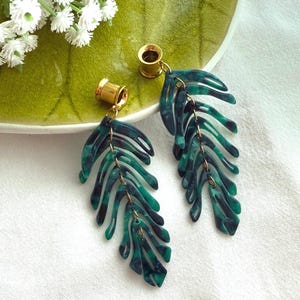 May include: A pair of dangle earrings featuring gold-toned ear tunnels attached to dark green and black marbled acrylic leaf-shaped pendants. The leaf designs have a cut-out, skeletal structure and are suspended from small metal jump rings.