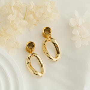 May include: A pair of gold-toned ear tunnels featuring dangling, irregular oval-shaped charms. Each tunnel has a smooth, polished metallic finish and is connected to a hollow, organic-shaped loop that hangs freely from the circular ear gauge base.