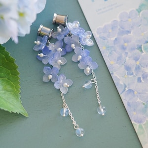May include: A pair of ear plugs featuring dangling silver chains adorned with translucent light blue hydrangea flower petals and small white beads. Each plug has a metallic silver base and the floral design hangs from the center of the plug.