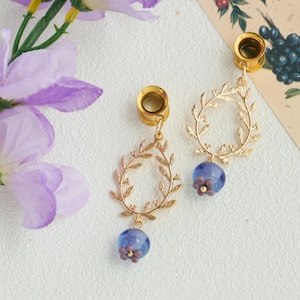 May include: Gold-colored ear gauges with a leaf-shaped design and a dangling blue bead with purple accents. The gauges are displayed on a white surface with purple flowers and botanical illustrations.