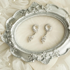 May include: A pair of silver-toned drop earrings featuring a circular stud with a central clear stone surrounded by smaller crystals, with a dangling leaf-shaped charm accented by marquise-cut stones, displayed inside an ornate silver frame.
