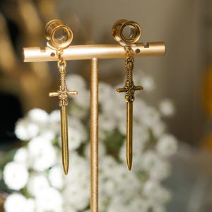 Pode incluir: Túneis de orelha dourados com pingentes de espada pendurados. Os túneis são circulares com uma borda alargada, e as espadas têm guardas intrincadas em forma de cruz. As joias são exibidas em um suporte dourado.