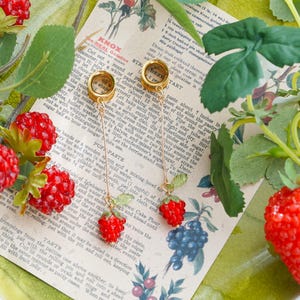 May include: A pair of gold-toned ear tunnels featuring dangling chains with small red strawberry charms. Each charm includes a tiny green leaf detail. The jewelry is displayed on a vintage-style paper recipe card with printed text and fruit illustrations.