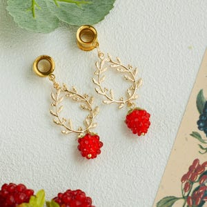 May include: Gold-toned earrings with a leaf-like design and a red, berry-shaped charm. The earrings are shown on a white surface, with a botanical illustration in the background.