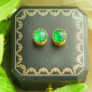 May include: A pair of green gemstone stud earrings with gold settings, displayed on a black jewelry box with gold decorative trim. The earrings are round and vibrant, set against a backdrop of green foliage.