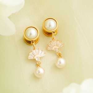 May include: A pair of gold-toned earrings with a pearl stud and a dangling fan-shaped charm adorned with clear crystals and a dangling pearl. The earrings are set against a soft, light green background.