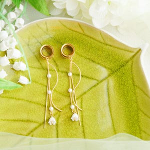 May include: Gold-coloured earrings with a delicate design. Each earring features a circular top and a flowing chain adorned with small white beads and a miniature white flower at the end. The earrings are displayed on a green leaf-shaped plate.