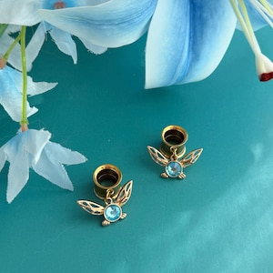 May include: A pair of gold-toned ear tunnels featuring dangling charms shaped like winged insects. Each charm has an intricate openwork wing design and a central round light blue crystal stone. The tunnels are circular with a polished finish.