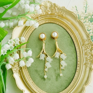 May include: Gold-toned earrings featuring a pearl stud and a cascading lily of the valley design. The earrings are displayed inside a gold ornate frame on a green background, with lily of the valley flowers in the foreground.