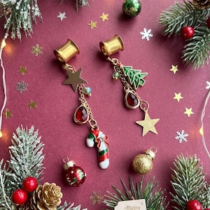 May include: A pair of gold-toned ear tunnels featuring dangling Christmas-themed charms. One tunnel has a gold star, a red teardrop crystal, and a candy cane. The other has a green Christmas tree, a red teardrop crystal, and a gold star.