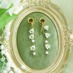 May include: Gold-coloured earrings with white lily of the valley flowers and faux pearls. The earrings hang from gold-coloured plugs, with a green leaf accent. The earrings are displayed on a green velvet background within a gold ornate frame.