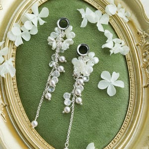 May include: A pair of dangle earrings featuring white floral details and hanging pearl-like beads. The earrings are displayed on a green velvet surface within a gold oval frame. White flower petals are scattered around the frame.