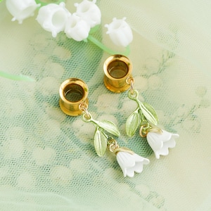 May include: Gold-coloured ear gauges with dangling lily of the valley charms. The charms feature white flower pendants with green leaves. The background is a light green fabric with white lily of the valley flowers.
