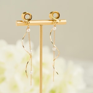 May include: Gold-toned earrings with a spiral design and dangling chains. Each earring features a small, round plug and is adorned with delicate white pearls. The earrings are displayed on a gold stand against a soft, blurred background.