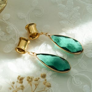 May include: A pair of gold-toned ear tunnels featuring dangling, faceted, teardrop-shaped green glass pendants. Each tunnel is connected to the gemstone by a small gold-coloured jump ring, creating a hanging design that catches the light.