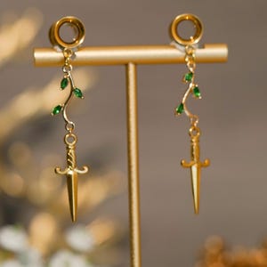 Pode incluir: Brincos dourados com pingente de adaga. Os brincos apresentam um aro dourado e um design pendurado com uma pequena adaga e detalhes de folhas verdes. Os brincos são exibidos em um suporte dourado.
