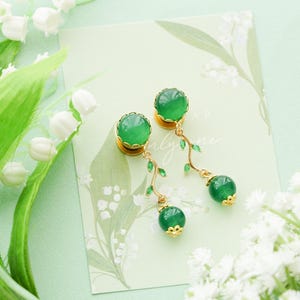 May include: A pair of green gemstone earrings with gold accents. The earrings feature a round green stone at the top and a dangling green bead. Delicate gold vines with tiny green leaves connect the beads. The earrings rest on a floral card.