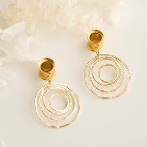 Könnte beinhalten: Ein Paar goldfarbene Ohrringe mit Spiralmuster. Die Ohrringe haben eine kreisförmige Form mit mehreren konzentrischen Ringen. Die Ohrringe hängen an einem goldfarbenen Pfosten. Der Hintergrund ist ein weiches Weiß.