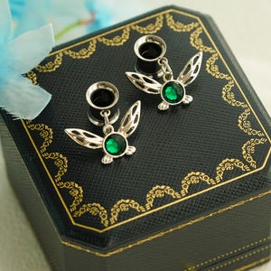 Può includere: Un paio di dilatatori per orecchie color argento con ciondoli a forma di ali pendenti, caratterizzati da una pietra preziosa circolare verde smeraldo al centro. I gioielli sono esposti su una scatola ottagonale nera con bordo dorato.
