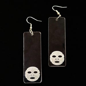 May include: A pair of clear acrylic earrings with a face mask design. The earrings are rectangular and have silver hooks.