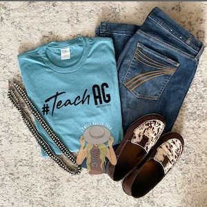 May include: A light blue t-shirt with the text "#TeachAG" in black. The shirt also features a cartoon illustration of a woman wearing a hat and a necklace. The text "Sweet Valley Tech" is below the illustration.