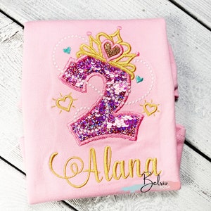 May include: Pink t-shirt with a gold crown and a sparkly pink number 2. The name "Alana" is embroidered in gold below the number. The shirt is folded in half.