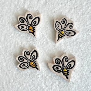 May include: Four ceramic bee-shaped embellishments. Each bee has a cream-colored body, black wing details, and a yellow and black striped abdomen. The bees are arranged on a white surface.