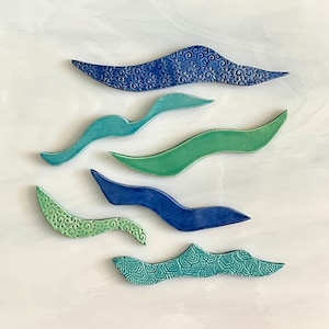 May include: An overhead shot displays six ceramic, wave-shaped pieces in shades of blue, green, and turquoise. Each piece has a unique textured pattern, with some featuring floral or geometric designs. The pieces are arranged on a white surface.