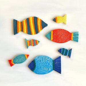 May include: A collection of six ceramic fish ornaments in various sizes and colors. The fish feature patterns including vertical stripes, swirls, and textured scales in shades of blue, orange, red, yellow, and teal with glossy finishes.