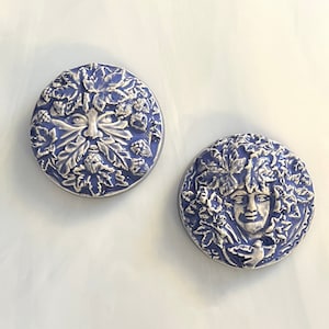 May include: Two round, blue ceramic Green Man face plaques. The faces are detailed with leaves and foliage, and the plaques have a raised, textured design. The faces are white and the background is blue.