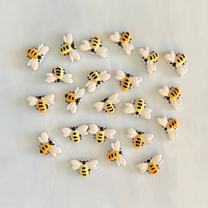 May include: A collection of small, decorative bee-shaped embellishments. Each bee features a yellow and black body with white wings. The bees are arranged on a light blue surface, creating a whimsical and charming aesthetic.