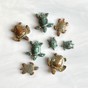 May include: A collection of eight ceramic turtle figurines in various sizes and colors. The turtles feature shades of brown, teal, and green with glossy finishes. The figurines are arranged on a white surface.