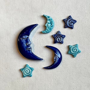 May include: A collection of ceramic celestial decorations, including crescent moons and star shapes. The moons feature serene faces and are in shades of blue and turquoise. The stars have a spiral design.