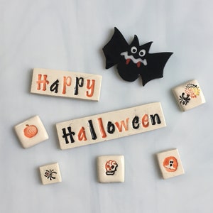 May include: Halloween-themed decorations including a black bat, and ceramic tiles with the words "Happy" and "Halloween". The tiles feature pumpkin, spider, skull, and ghost designs. The text and designs are in orange and black.