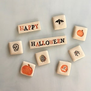 May include: A collection of Halloween-themed ceramic tiles. The tiles feature various designs, including a skull, pumpkin, bat, spider, and the words "HAPPY" and "HALLOWEEN" in orange and black lettering. The tiles are arranged on a light blue surface.