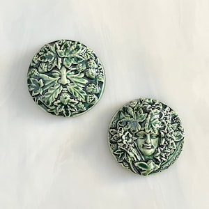 May include: Two green ceramic Green Man face plaques. One plaque features a face surrounded by leaves and berries. The other plaque shows a face surrounded by leaves and flowers. Both plaques are round and have a glossy finish.