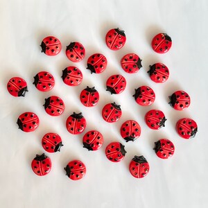 May include: A collection of red ladybug-shaped decorative objects with black accents. Each ladybug has a glossy finish and small black dots on its red shell. The ladybugs are scattered on a white surface.