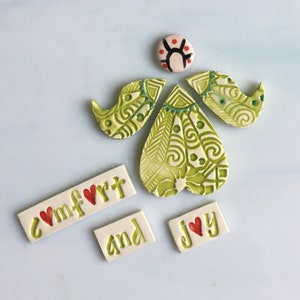 May include: A collection of ceramic pieces in a light green and white color scheme. The pieces include an angel-like figure with decorative patterns, and rectangular tiles spelling out "comfort and joy" with red heart accents. The angel has a round head with a black and red design.