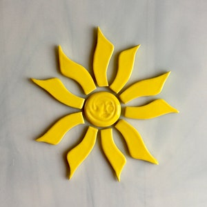 May include: A bright yellow sun-shaped decorative tile. The sun has a smiling face in the center and curved rays extending outwards. The tile is made of a glossy material and is set against a light background.